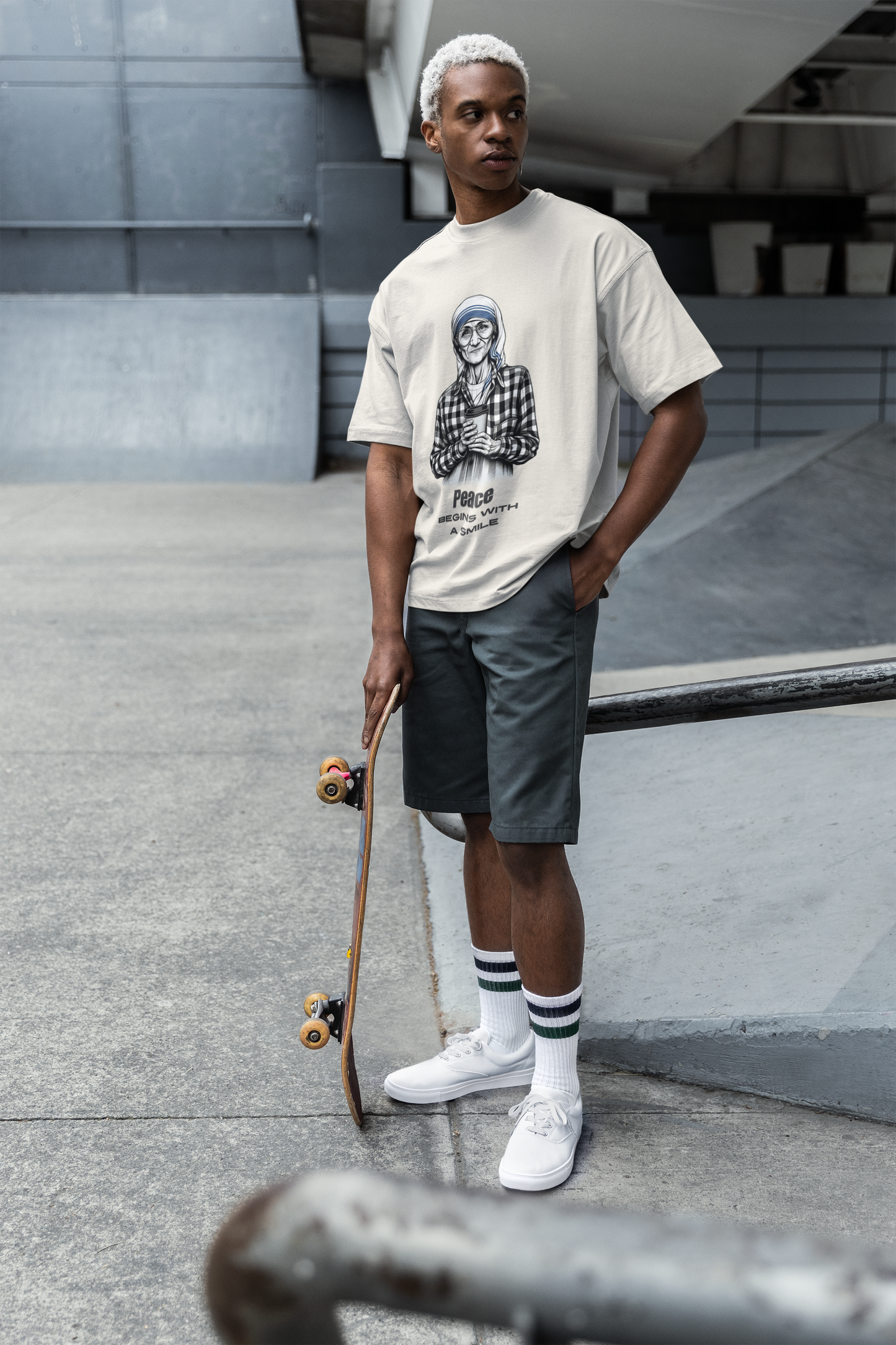 man wearing an oversized Mother Teresa t-shirt in faded bone color, holding a skateboard. The t-shirt features a detailed sketch of Mother Teresa with the quote "Peace Begins With a Smile." The scene is set in an urban environment, showcasing a casual and stylish look perfect for everyday wear.