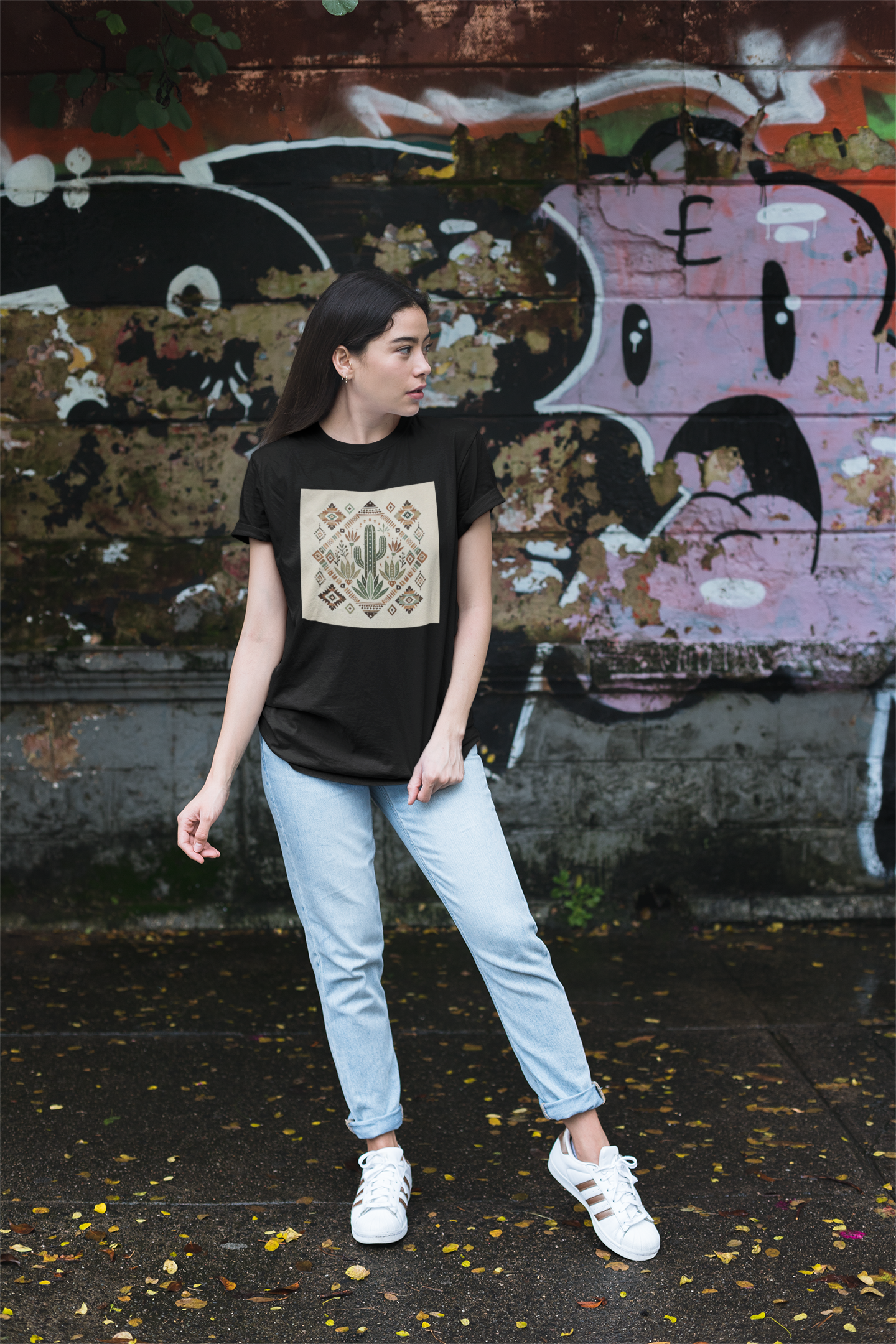 Woman wearing a black t-shirt with a cactus and Aztec pattern graphic, standing in front of a graffiti wall.